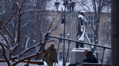 засніжений сквер із пам’ятником Степану Бандері, поруч майорить синьо-жовтий прапор. На передньому плані видно зламані металеві конструкції та гілки, а двоє чоловіків у зимовому одязі прибирають територію