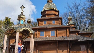 Дерев'яний храм із золотими банями. Біля храму розташована фігура Матері Божої у червоно-блакитному вбранні. Довкола святині ростуть високі зелені дерева. Фото з сайту 