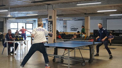 Двоє чоловіків грають у настільний теніс у просторі Urban Camp. Довкола — інші відвідувачі: хтось відпочиває на диванах, а хтось спостерігає за грою чи займається своїми справами.