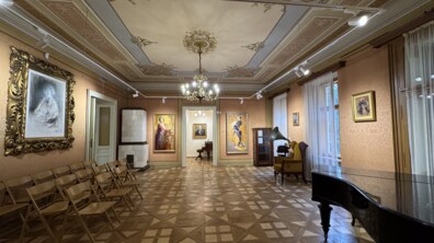 Простора світла зала з високою, багато прикрашеною стелею та дерев’яною підлогою з чітким геометричним візерунком. Уздовж стін висять картини в золотих рамах, ліворуч стоять ряди стільців, а праворуч — чорний рояль і старовинний грамофон.