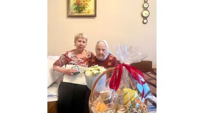 Столітня ювілярка Лідія Булідорова та її родичка сидять у кімнаті; жінка тримає в руках вітальний лист та памʼятну монету; перед ними стоїть кошик з фруктами, а позаду на стіні висять картина та годинник