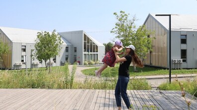 На фото мама піднімає на руках маленьку дівчинку. На задньому плані — сучасні будівлі Центру матері та дитини “Незламні матусі”, зелені насадження, дерева та доріжки.