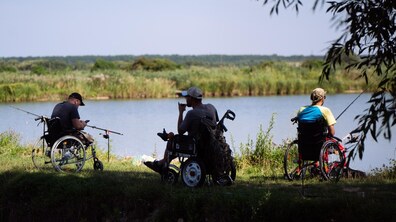 Троє ветеранів на кріслах колісних ловлять рибу на березі озера. Вони відпочивають, насолоджуючись природою та краєвидом. На задньому плані видно спокійну воду, очерет і зелену рослинність, а праворуч зверху — гілки дерева.