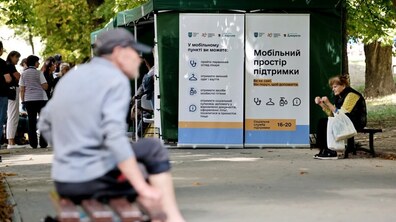 «Мобільний простір підтримки», облаштований у зелених наметах біля Порохової вежі. На передньому плані видно чоловіка, який сидить на лавці спиною до камери. Позаду нього розташовані намети, біля яких перебувають люди. На інформаційних стендах зазначено, що тут можна отримати первинну медичну допомогу, засоби особистої гігієни, змінний одяг і взуття, а також соціальну підтримку. Праворуч жінка сидить на лавці з пакетом у руках.