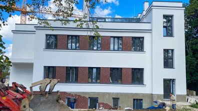 Будинок підтриманого проживання на етапі завершальних будівельних робіт. Це сучасна триповерхова будівля з пласким дахом, фасад поєднує білі стіни зі вставками з червоної цегли та великими темними вікнами. На передньому плані видно будівельну техніку й робітників, які виконують роботи біля будівлі. Навколо є дерева, а в небі – ясна погода