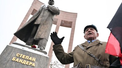 На передньому плані стоїть чоловік у військовому однострої, який звертається до присутніх. Поруч видно червоно-чорний прапор. Позаду пам’ятник Степану Бандері