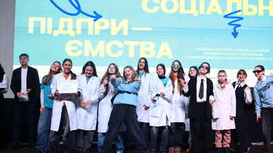 Учні у різних костюмах стоять на сцені та посміхаються. Дівчинка тримає у руках сертифікат. Позаду на екрані напис великими жовтими літерами “Шкільні соціальні підприємства”.