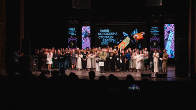 На фото — велика група людей стоїть на сцені під час офіційної події. Перед ними мікрофон і трибуна, а позаду — великий екран із написом «Львів — Молодіжна столиця Європи 2025».