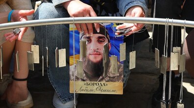 «Місце надії» у центрі Львова: на крупному кадрі видно як іменні жетони висять на металевій конструкції, а поруч є люди. По центру композиції жінка тримає портрет військового з позивним «Борода», де написано: «Любимо. Чекаємо».