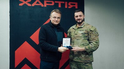 Мер Львова Андрій Садовий та командир корпусу «Хартія» Ігор Оболенський Ігор разом тримають відкритий футляр із міською відзнакою – нагрудним знаком «Залізний Лев» на тлі великого чорно-червоного банера з символікою та написом «Корпус Хартія».