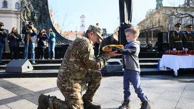 На площі перед пам’ятником військовослужбовець у камуфляжі, ставши на одне коліно, тримає разом із хлопчиком складений синьо-жовтий прапор. Позаду стоять фотографи, глядачі та військові почесної варти