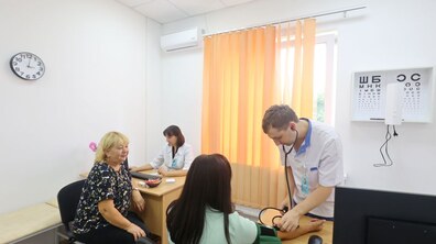 У медичному кабінеті лікар в білому халаті вимірює артеріальний тиск жінці, яка сидить за столом. Поруч на стільці сидить інша жінка, яка дивиться у бік пацієнтки. На задньому плані інша медична працівниця працює за столом, а на стіні видно таблицю для перевірки зору.