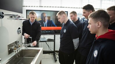 Чоловік середнього віку показує групі юнаків, як користуватися обладнанням біля металевої мийки з краном у навчальному класі. Він тримає в руках деталь і пояснює процес. Поруч стоять кілька хлопців у темному робочому одязі, уважно спостерігають і слухають. На задньому плані видно великий екран із презентацією, а також навчальне обладнання.