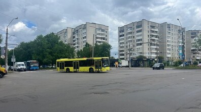 Перехрестя вул. Широка і вул. Суботівська з жовтим автобусом у центрі. На фоні розташовані сірі багатоповерхівки, зелені дерева та невеликі магазини. Небо хмарне, асфальт на передньому плані нерівний.