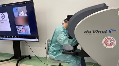 Медичний працівник у захисному одязі працює за роботизованою хірургічною системою DaVinci. Ліворуч розташований великий екран із відеоконференцією, де триває трансляція навчальної операції.