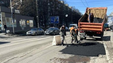 Ремонтна бригада ремонтує дорожнє покриття на міській вулиці. Біля узбіччя стоїть вантажівка з відкритим кузовом, наповненим гарячим асфальтом, а один із працівників у кузові подає матеріал донизу. Двоє робітників у помаранчевих жилетах лопатами розрівнюють свіжий чорний асфальт на ямі на дорозі, поруч стоїть дорожній конус, що позначає місце ремонту. На задньому плані видно автомобілі, будівлі та дерева.