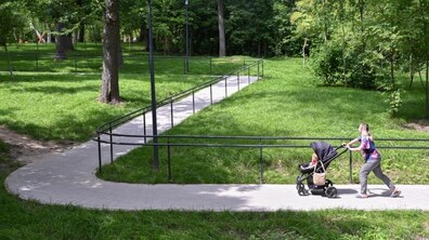 Мати з дитиною у візочку гуляє інклюзивною доріжкою Замарстинівського парку. Довкола зелена трава та дерева
