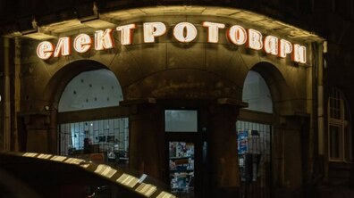 Нічний вигляд крамниці, розташованої на першому поверсі старовинного будинку з арковими вікнами. Над входом яскраво світиться автентична вивіска неоновими або люмінесцентними літерами «електротовари», що виділяється на тлі темного фасаду