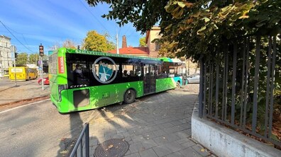 Великий зелений міський автобус повертає з проспекту Чорновола на площу 700-річчя Львова. Праворуч є металева огорожа та дерева із зеленим листям.