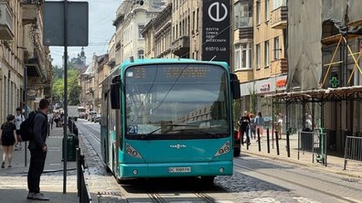 Бірюзовий автобус №21 на міській вулиці. З обох боків видно будинки та перехожих.