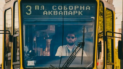 Крізь лобове скло жовтого трамвая видно чоловіка-водія у сонцезахисних окулярах та вишиванці, який сидить у кабіні. Над склом на електронному табло світиться маршрут №3: «пл. Соборна — Аквапарк»