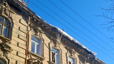 Верхня частину старого багатоповерхового будинку з кам’яним фасадом. На даху лежить шар снігу що може впасти вниз. З одного з верхніх вікон чоловік висунув довгу палицю і нею намагаються збити сніг та бурульки з даху.