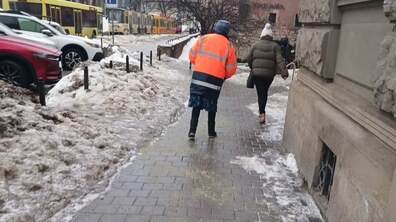 Двірничка, вдягнена в яскраву помаранчеву куртку зі світловідбивними смугами, посипає піском вкритий льодом тротуар, щоб зробити поверхню менш слизькою. Попереду праворуч іде пішохід. Обабіч тротуару лежить сніг, поруч припарковані автомобілі, а далі видно жовті трамваї.
