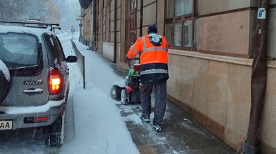 чоловік двірник, одягнутий у спецодяг, чистить від снігу тротуар із допомогою ручної техніки. Справа від нього будинок світлокоричневого кольору, зліва – припарковане авто