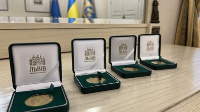 Чотири відкриті темні футляри з пам’ятними монетами Короля Данила. Усередині кожної коробочки на білій шовковій кришці зображено логотип Львова, а на тлі у дзеркалі відображені прапори України, Європейського Союзу та Львова.