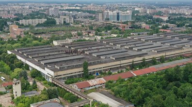 Панорамний вигляд з висоти на великий промисловий комплекс Львівського автобусного заводу та навколишню територію. У центрі кадру — довгі виробничі корпуси, що свідчать про індустріальне призначення будівель. На передньому плані видно зелені насадження, старі господарські будівлі та естакаду, яка з’єднує частини комплексу. На тлі — сучасні житлові квартали Львова, багатоповерхівки та міська забудова, що підкреслює контраст між індустріальним минулим території й активним розвитком міста навколо.