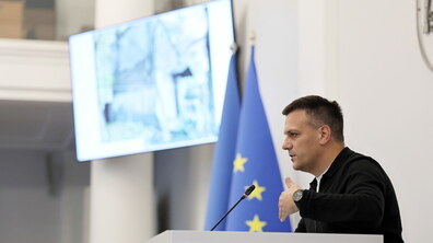 Депутат Юрій Лукашевський стоїть за трибуною у сесійній залі. Біля нього видніються прапори. На стіні висить великий телевізор, на якому транслюють фото