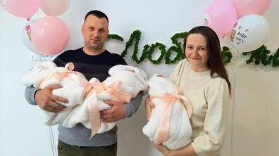 Подружжя Руслан та Наталія Кукурудзи усміхаються і тримають на руках новонароджених донечок Анну, Софію та Вікторію, загорнутих у м’які білі пледи та перев’язані ніжно-рожевими стрічками. Позаду видно святкові повітряні кульки.