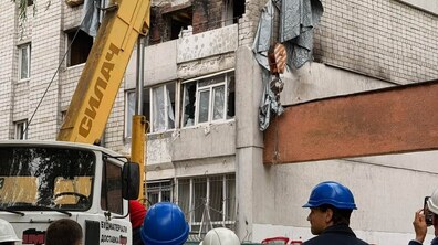 Делегація із декількох людей у захисних будівельних касках дивляться на зруйновану багатоповерхівку, де вибиті вікна і знищена стіна будинку внаслідок російської атаки