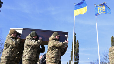 Військовослужбовці у формі несуть труну на військове кладовище. На фоні чистого блакитного неба майорять Прапор України та Прапор Львова