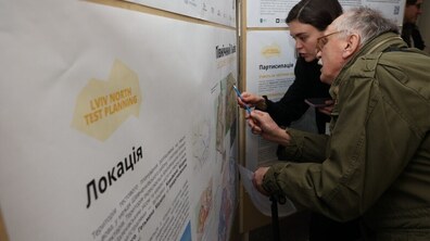 Презентація матеріалів про розвиток північної частини Львова: на передньому плані двоє учасників уважно розглядають інформаційні стенди з картами, схемами та текстами. На задньому плані розміщені інші інформаційні плакати, присвячені плануванню території та участі мешканців у формуванні майбутнього північного Львова.
