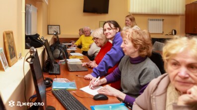 Навчальне заняття в комп’ютерному класі для людей старшого віку. Викладачка допомагає учасникам працювати за комп’ютерами та ноутбуками: усі уважно слухають, роблять нотатки та навчаються цифровим навичкам.
