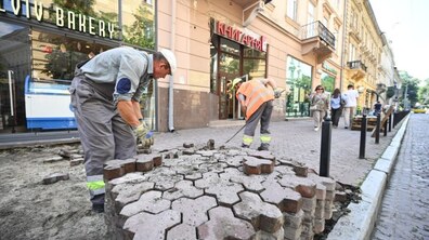 Ремонтні роботи з облаштування доступних входів на вул. Січових Стрільців. На передньому плані двоє працівників у робочому одязі: один у сірій формі та кепці нахилився і вручну викладає фігурну бруківку, інший у помаранчевому жилеті розрівнює основу для мощення.