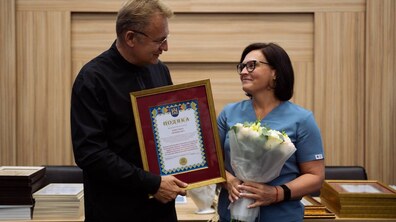 міський голова Львова вручає подяку медичній працівниці. Він тримає у руках велику рамку з грамотою, а жінка в блакитному медичному костюмі — букет білих квітів. Вони усміхаються й дивляться одне на одного.