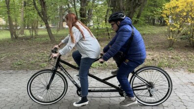 Двоє людей їдуть на велотандемі парковою доріжкою. Попереду — дівчина, супроводжуюча, з довгим рудим волоссям у світлій сорочці та джинсах, яка керує велосипедом. Позаду — хлопець у синій куртці та шоломі, який разом із нею крутить педалі
