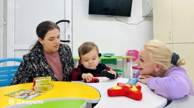 Маленька дитина сидить за столом і грається іграшковими машинками. Поруч із нею дві жінки: одна уважно спостерігає, а інша усміхається, підтримуючи малюка. Вони займаються у світлій ігровій кімнаті центру з яскравими меблями та іграшками. Атмосфера виглядає теплою та дружньою.