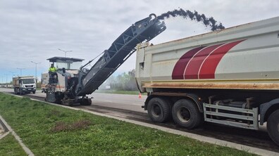 Фрезерування дорожнього покриття на вул. Стрийській. Уздовж дороги стоїть велика дорожня фреза, яка знімає верхній шар асфальту. Чорна подрібнена маса через довгий транспортер пересипається у кузов вантажівки, що їде поруч. З ліва видно смугу трави на узбіччі.
