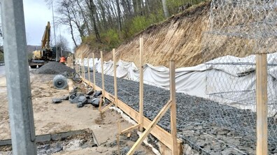 Будівельний майданчик біля підніжжя крутого земляного схилу. Вздовж схилу встановлюють габіони — це великі сітчасті ящики з товстого металевого дроту, наповнені сірим камінням. На передньому плані видно один такий довгий ярус, уже заповнений щебенем. Між камінням і самим земляним схилом прокладено біле полотно (геотекстиль)