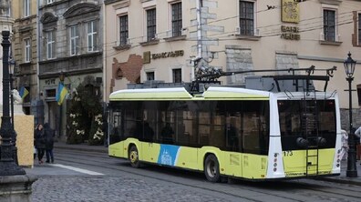 Сучасний жовтий тролейбус марки «Електрон» рухається міською вулицею на тлі кам’яниць . Його штанги опущені й зафіксовані на даху, що підкреслює рух машини на автономному ходу.