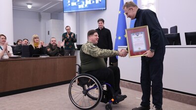 Міський голова вручає подяку. Ветеран у військовій формі на кріслі колісному тисне руку меру. Посадовці аплодують у Великій сесійній залі Львівської міської ради