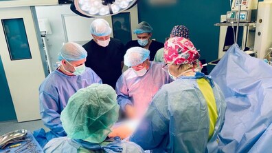 Хірургічна операція: п’ятеро медичних працівників у стерильних захисних костюмах, масках та шапочках працюють в операційній під яскравим операційним світлом. Поруч є різноманітні хірургічні інструменти, що розкладені на стерильному покритті, а також медичне обладнання.