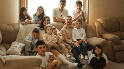 Велика родина Андрія та Олени Видюків у затишній вітальні: дорослі, підлітки та маленькі діти сидять на дивані й навколо нього, а на передньому плані хлопець тримає руде кошеня. Всі у гарному настрої, дехто усміхається, дивлячись в камеру.