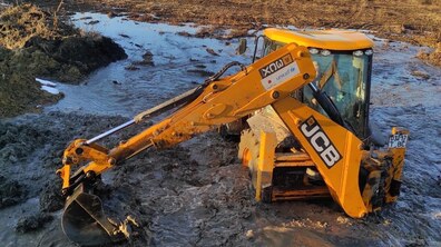 Жовтий екскаватор працює у великій калюжі води на відкритій ділянці місцевості. Техніка частково занурена в рідкий мул — колеса і нижня частина корпусу у воді та болоті. Передній ківш опущений у багнюку й зачіпає мокру землю. На задньому плані — кілька приватних будинків із жовтими та червоними фасадами, господарські споруди та дерева.