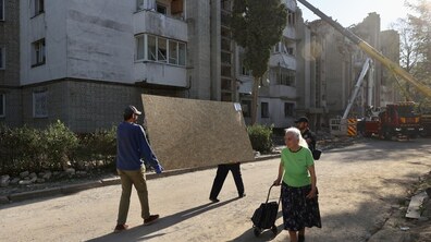 На передньому плані двоє чоловіків несуть велику дерев’яну плиту для закриття пошкоджених вікон. Поруч дорогою йде літня жінка з невеликим візком для покупок. Частина фасаду будинку та верхніх поверхів пошкоджена, вибиті вікна, на місці працює великогабаритний кран