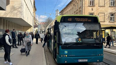 Зупинка громадського транспорту в центрі Львова. Біля платформи стоїть низькопідлоговий автобус №48, а на електронному табло вказаний його маршрут. На тротуарі поруч чекають пасажири: частина людей стоїть біля зупинки, а літня жінка з візком для покупок іде вздовж платформи.