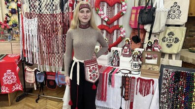 Юна дівчина з довгим світлим волоссям стоїть у кімнаті, заповненій виробами ручної роботи. На ній бежевий гольф, чорні штани та вишитий червоно-чорний пояс із кишенею. Навколо неї — виставка: плетені сітки (макраме), сумки з вишивкою, ляльки-мотанки та великі червоні декоративні серця.